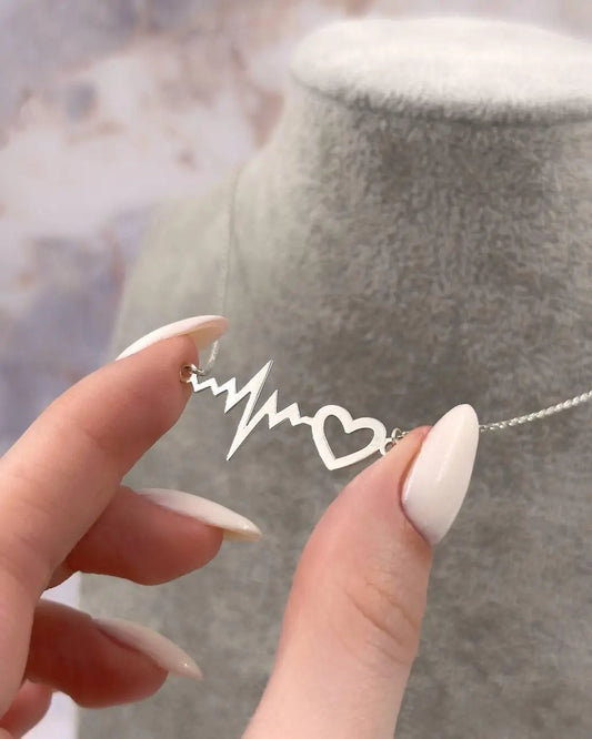 Elegant silver bracelet with a heart and heartbeat charm, symbolizing love and vitality. Displayed on a minimalist white ceramic dish, the bracelet shines against a soft purple striped background. Green leaves and delicate white flowers add a natural, romantic accent. The subtle “Apt. 317” text in the corner evokes a boutique aesthetic, perfect for gifting or everyday wear. Ideal for medical professionals, loved ones, or anyone celebrating life’s pulse.